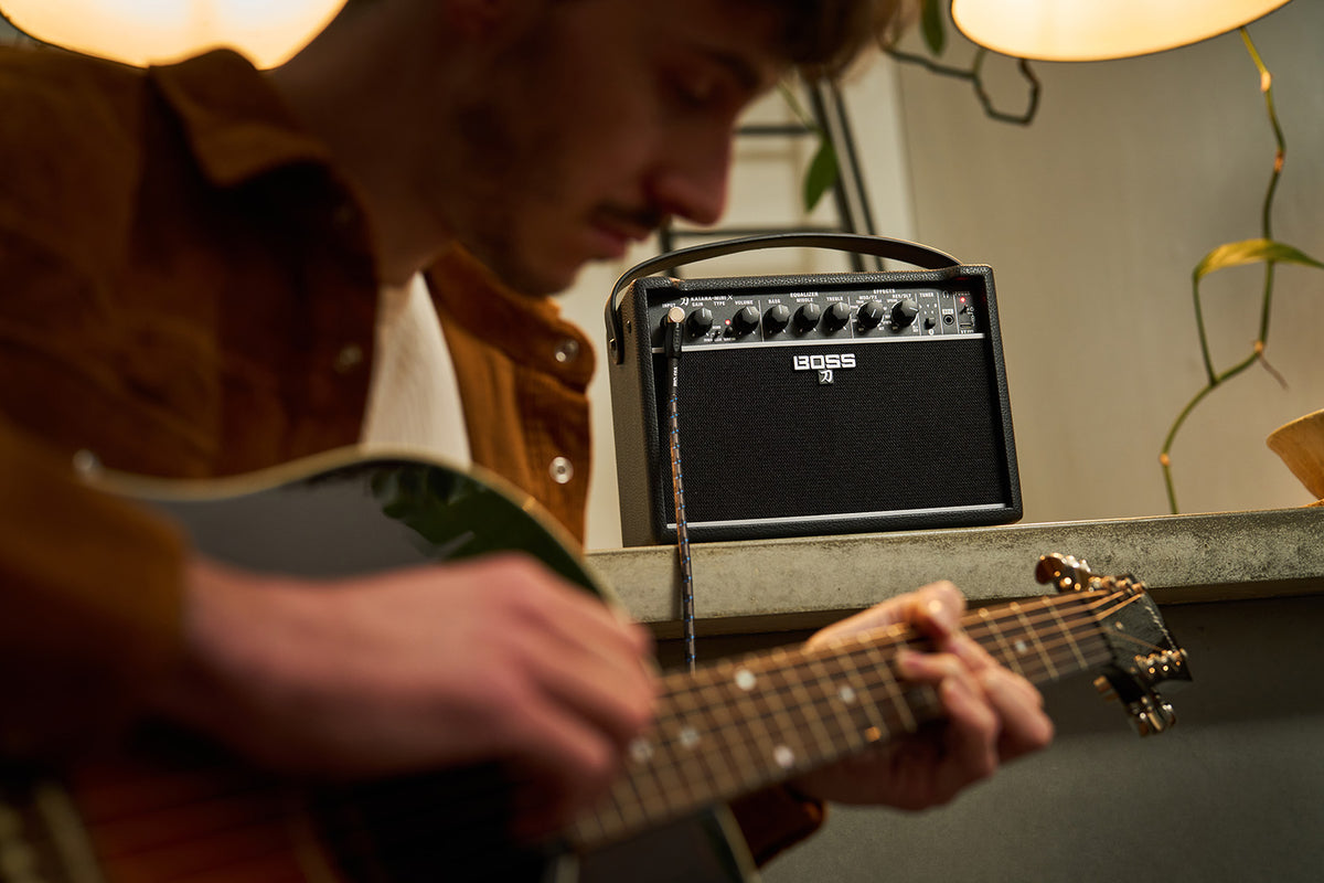 Boss Katana Mini X black, front view with guitarist playing acoustic guitar in background